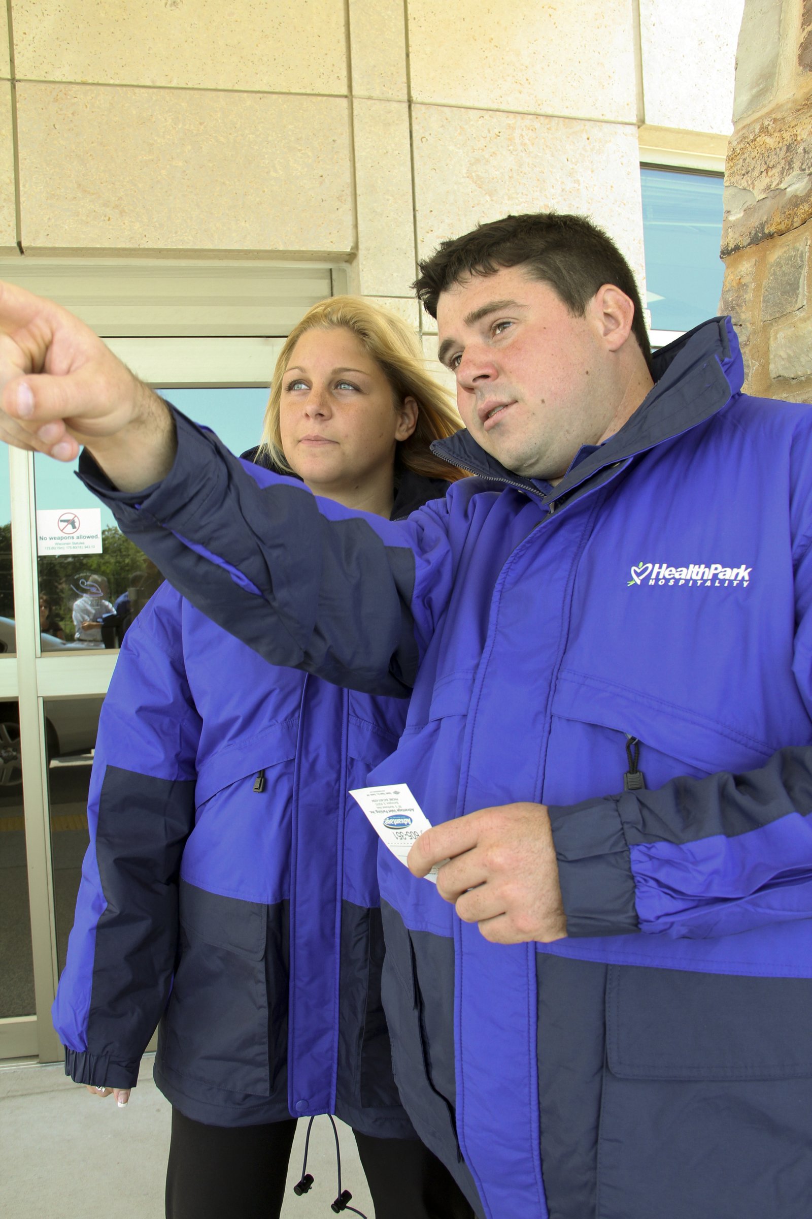Two HealthPark Hospitality staff members in blue parkas outside a building, with one pointing and holding a ticket